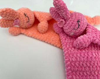 May include: Two crocheted bunny plushies, one pink and one peach, are lying together on a blanket. The bunnies have embroidered eyes and noses.