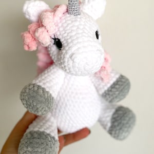 May include: A white crocheted unicorn stuffed animal with a pink mane and a silver horn. The unicorn has grey felt hooves.