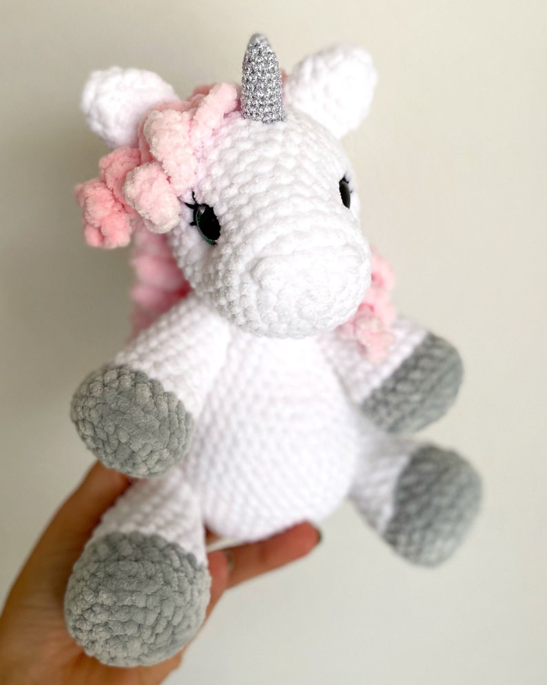 May include: A white crocheted unicorn stuffed animal with a pink mane and a silver horn. The unicorn has grey felt hooves.