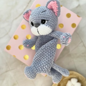 May include: A handmade gray crochet mouse toy with pink inner ears and paw pads. The plush toy is lying on a pink box with gold polka dots. The mouse has a white muzzle and black eyes.