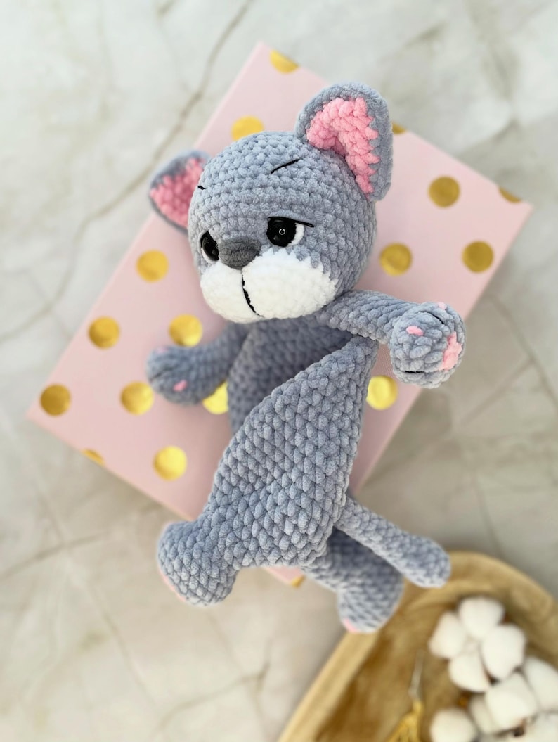 May include: A handmade gray crochet mouse toy with pink inner ears and paw pads. The plush toy is lying on a pink box with gold polka dots. The mouse has a white muzzle and black eyes.