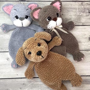 Op de afbeelding: Drie handgemaakte pluche speelgoed: een grijze muis met roze oren, een bruine kat met een witte strik en een bruine hond. Elk zacht speelgoed heeft zwarte ogen en is gemaakt van een getextureerd, pluche materiaal. Het speelgoed ligt op een lichtgekleurd houten oppervlak.
