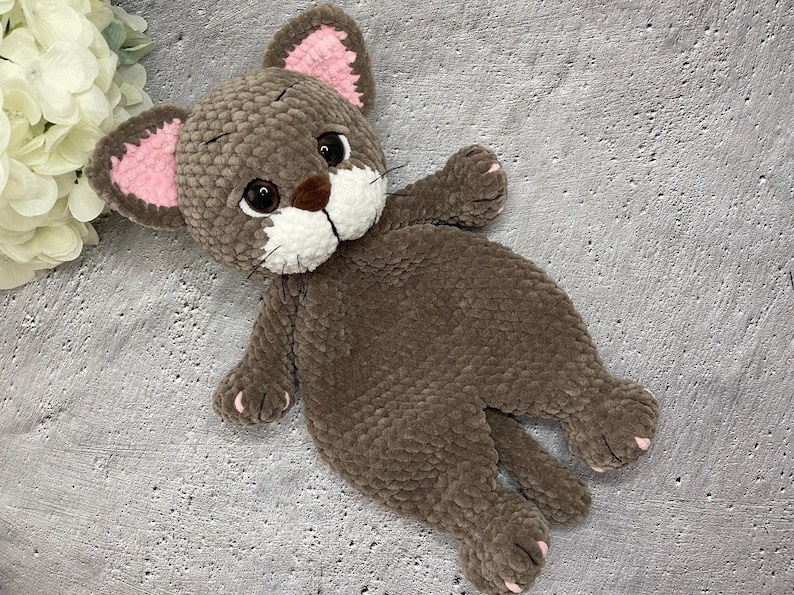 May include: A handmade brown plush cat toy with pink inner ears, white muzzle, and black eyes. The cat has a soft, textured appearance and is lying down. The toy is a cuddly, child-friendly item.