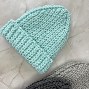 May include: A light blue knitted beanie hat with a folded brim. The hat is made of a chunky knit material. Two other knitted hats in gray and dark gray are partially visible.