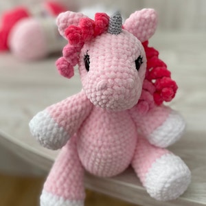 May include: A pink crocheted unicorn stuffed animal with a white horn and pink mane. The unicorn is sitting on a white surface.