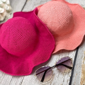 Puede incluir: Dos sombreros de sol de ganchillo en tonos rosa y melocotón, con alas anchas y copas redondeadas. Un par de gafas de sol con montura dorada y lentes degradadas descansa cerca. Los sombreros están sobre una superficie de madera clara y envejecida.
