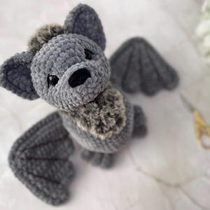 May include: A handcrafted grey plush bat with large ears and wings. The bat has a fluffy, textured body and a patch of brown fur on its chest. The bat's eyes are black, and it has a black nose. The wings are a darker shade of grey.