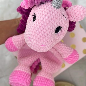 May include: A handmade, plush unicorn in shades of pink and magenta. The unicorn has a silver horn, black eyes, and a fluffy mane and tail. The soft toy is made of a textured yarn, giving it a cozy appearance.