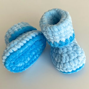 May include: A pair of baby booties made of blue fuzzy yarn. The booties are crocheted and have a light blue and dark blue ombre effect.