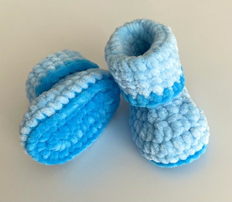 May include: A pair of baby booties made of blue fuzzy yarn. The booties are crocheted and have a light blue and dark blue ombre effect.