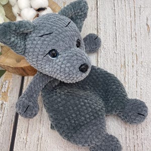 May include: A grey crochet dog toy with black eyes and nose. The dog has floppy ears and is lying down. The toy is made of soft, textured yarn. The dog is on a wooden surface.
