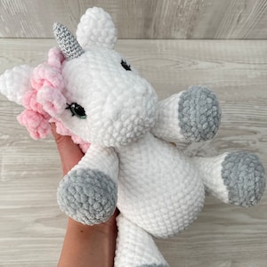 May include: A white crocheted unicorn stuffed animal with a pink mane and a silver horn. The unicorn has gray crocheted accents on its hooves and tail.