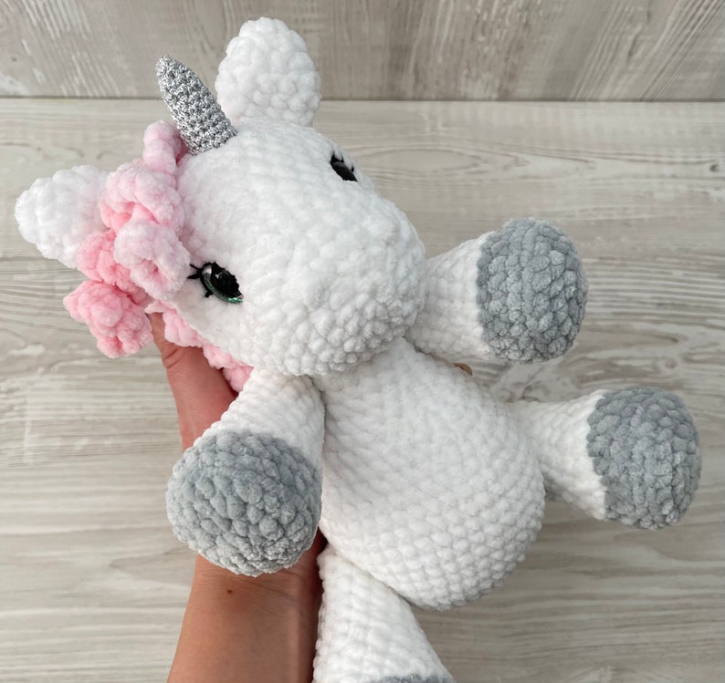 May include: A white crocheted unicorn stuffed animal with a pink mane and a silver horn. The unicorn has gray crocheted accents on its hooves and tail.