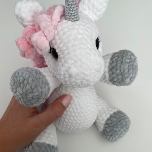 May include: A white crocheted unicorn stuffed animal with a pink mane and a silver horn. The unicorn has gray crocheted hooves.