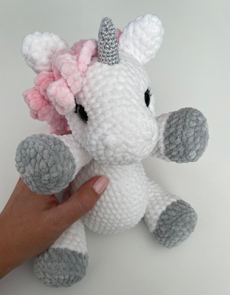May include: A white crocheted unicorn stuffed animal with a pink mane and a silver horn. The unicorn has gray crocheted hooves.