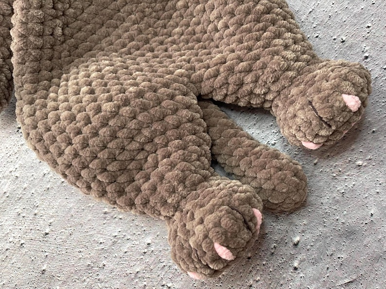 May include: A close-up of a brown, chunky knit blanket with two paws visible. The paws have pink accents on the pads and claws. The blanket is made of a soft, plush material.
