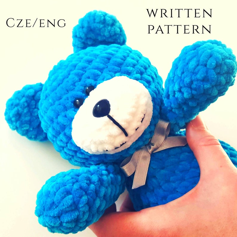 Puede incluir: Un oso de peluche azul tejido a crochet con detalles blancos en la cara y una cinta plateada. El oso est&aacute; siendo sostenido por una mano.