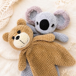 May include: A crochet security blanket featuring a tan bear head with a cream muzzle and black eyes, layered over a light gray koala head with large fuzzy ears and a black nose. The soft, plush yarn construction creates a textured, cuddly appearance.