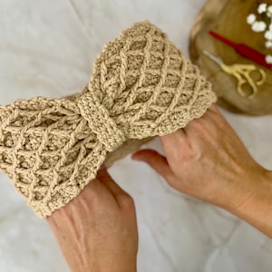 May include: A beige, hand-crocheted headband with a bow design. The headband features a textured, open weave pattern and is made of a soft, natural-coloured yarn. The bow is centered and adds a decorative element to the headwear.