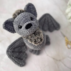 May include: A handmade gray crochet bat plush toy with large ears and wings. The bat has a fluffy chest and a black nose and eyes. The wings are spread out, and the toy is sitting on a white surface.