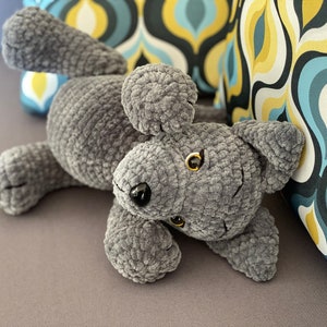 Pu&ograve; includere: Un giocattolo di peluche per cani grigio all'uncinetto con occhi neri e naso nero. Il cane &egrave; sdraiato sulla schiena con le zampe in aria.