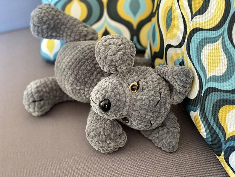 Pu&ograve; includere: Un giocattolo di peluche per cani grigio all'uncinetto con occhi neri e naso nero. Il cane &egrave; sdraiato sulla schiena con le zampe in aria.