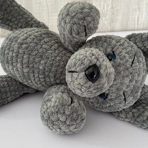 Pu&ograve; includere: Un giocattolo di peluche a forma di cane grigio lavorato a uncinetto con occhi azzurri e naso nero. Il cane &egrave; sdraiato di lato con la testa girata.