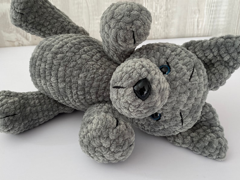 Pu&ograve; includere: Un giocattolo di peluche a forma di cane grigio lavorato a uncinetto con occhi azzurri e naso nero. Il cane &egrave; sdraiato di lato con la testa girata.