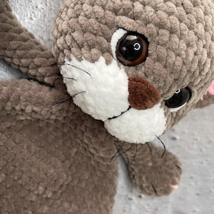May include: A handmade plush cat toy in shades of brown and gray. The cat has large, dark eyes, a white muzzle, and black whiskers. The toy is made of soft, textured yarn, and the overall design is cute and cuddly.
