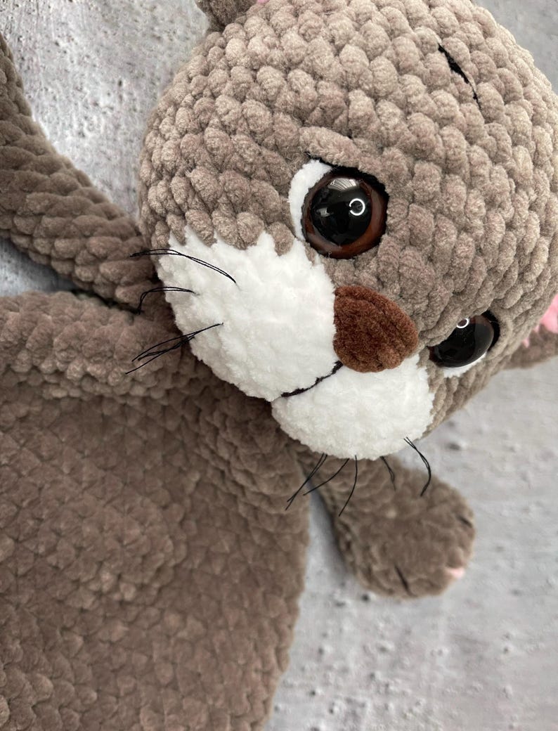 May include: A handmade plush cat toy in shades of brown and gray. The cat has large, dark eyes, a white muzzle, and black whiskers. The toy is made of soft, textured yarn, and the overall design is cute and cuddly.