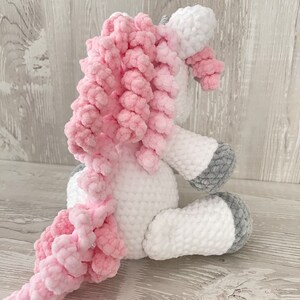 May include: A white crocheted unicorn with a pink curly mane and tail. The unicorn is sitting on a white wooden surface.