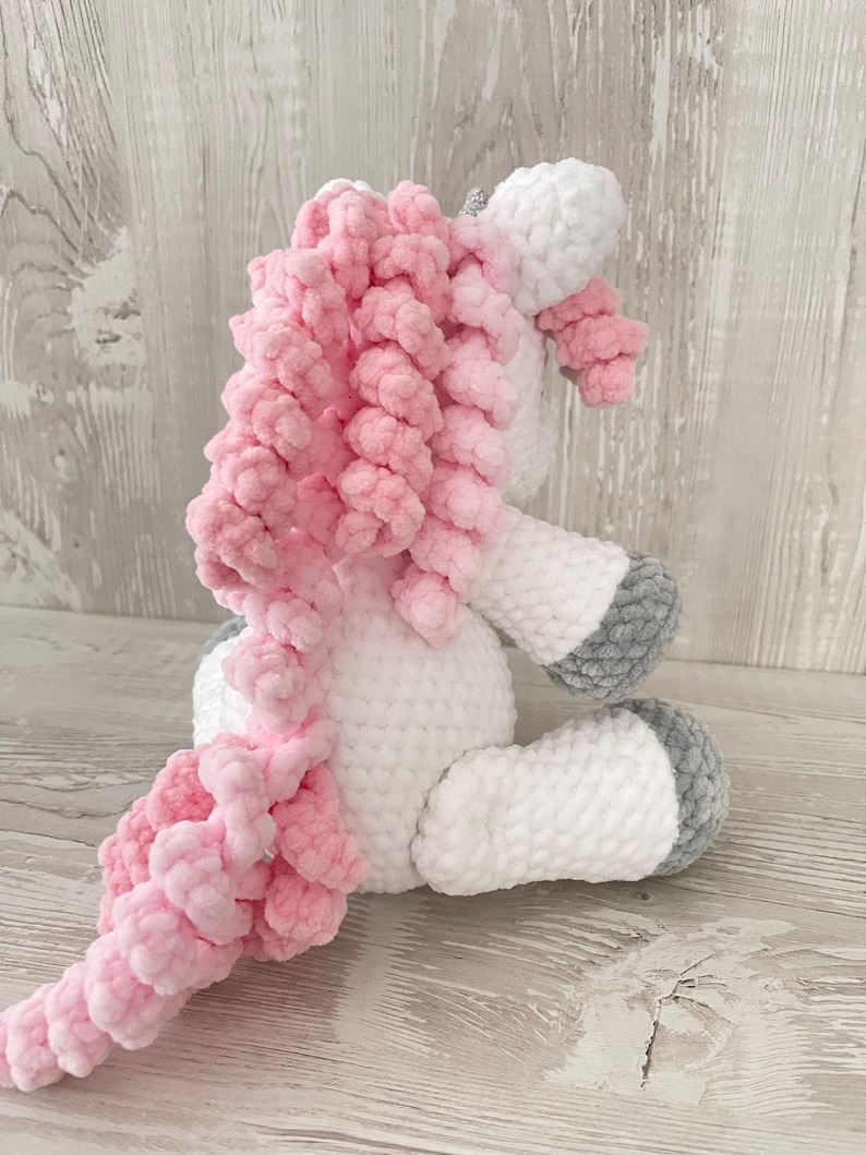 May include: A white crocheted unicorn with a pink curly mane and tail. The unicorn is sitting on a white wooden surface.