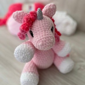 May include: A pink crocheted unicorn with a silver horn and pink mane. The unicorn is sitting on a light wood surface.