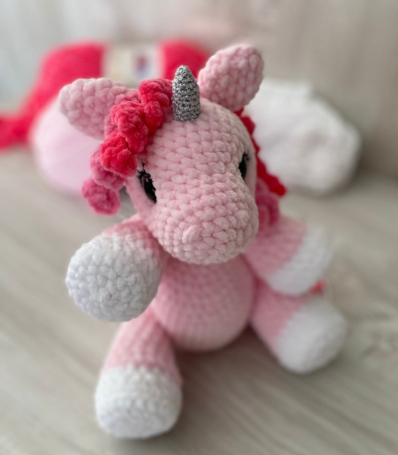 May include: A pink crocheted unicorn with a silver horn and pink mane. The unicorn is sitting on a light wood surface.