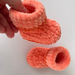 May include: A pair of coral-colored crocheted baby booties with a contrasting orange stripe around the ankle.