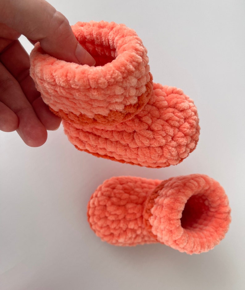 May include: A pair of coral-colored crocheted baby booties with a contrasting orange stripe around the ankle.