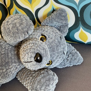 Pu&ograve; includere: Un animale di peluche grigio a forma di cane, lavorato a uncinetto, sdraiato su un divano grigio. Il cane ha gli occhi neri e il naso nero.