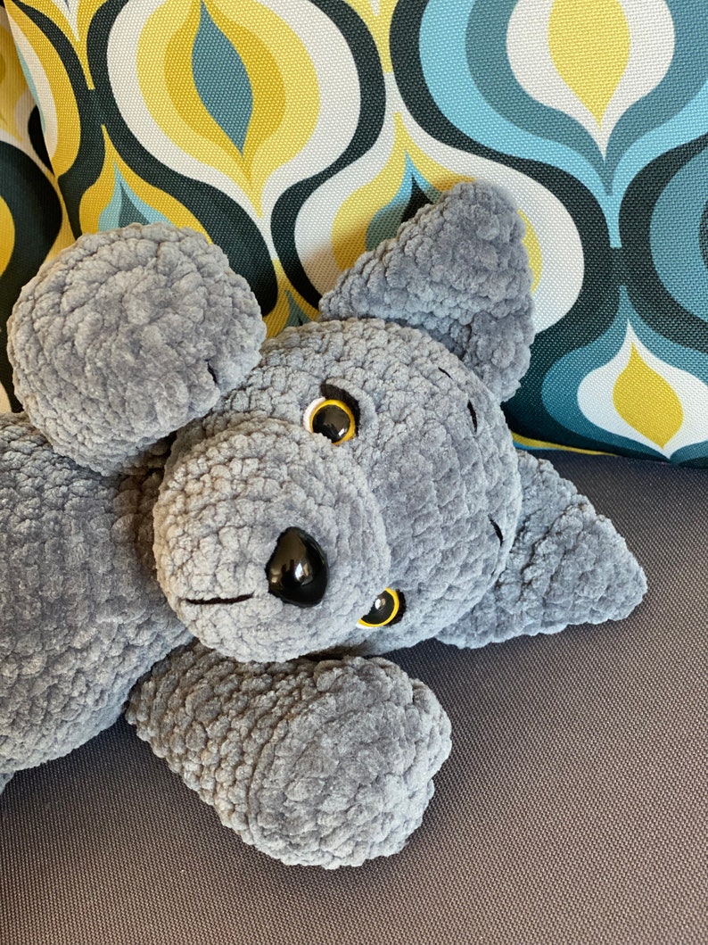 Pu&ograve; includere: Un animale di peluche grigio a forma di cane, lavorato a uncinetto, sdraiato su un divano grigio. Il cane ha gli occhi neri e il naso nero.