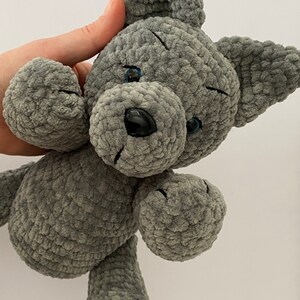 Pu&ograve; includere: Un cane di peluche grigio lavorato a uncinetto con occhi blu e naso nero. Il cane &egrave; seduto con le zampe allungate.