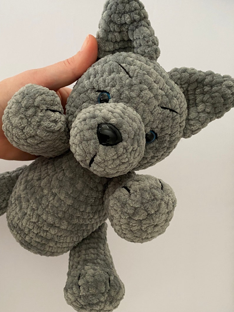 Pu&ograve; includere: Un cane di peluche grigio lavorato a uncinetto con occhi blu e naso nero. Il cane &egrave; seduto con le zampe allungate.