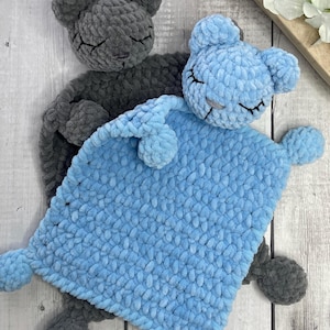 Pode incluir: Duas mantas de segurança para bebés feitas à mão em croché. Uma é cinzenta, a outra azul clara. Cada uma tem uma cara de urso com os olhos fechados e orelhas pequenas. As mantas são macias e felpudas, perfeitas para um bebé.