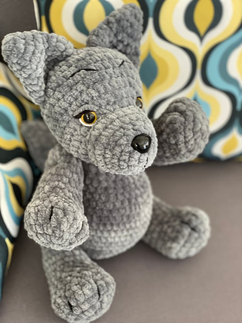 Pu&ograve; includere: Un cane di peluche grigio lavorato a uncinetto con occhi neri e naso giallo. Il cane &egrave; seduto su un divano grigio.