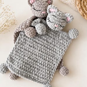 May include: Two handmade crochet baby security blankets in the shape of sleeping cats. The blankets are gray and light brown, with pink accents on the ears and noses. The cats have closed eyes and are made of soft, textured yarn.