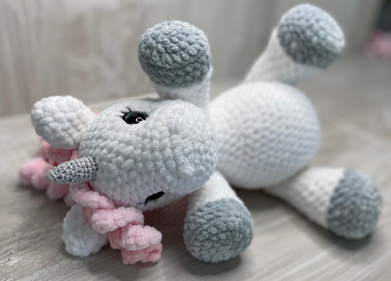May include: A white crocheted unicorn plush toy with a pink mane and tail. The unicorn has a silver horn and gray hooves.