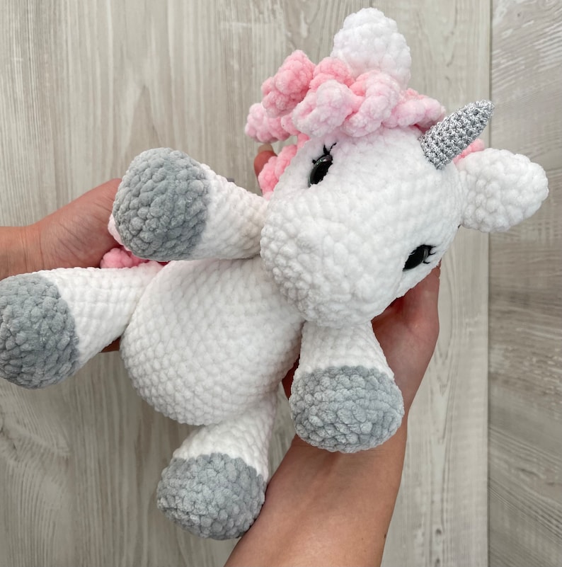 May include: A white crocheted unicorn stuffed animal with pink yarn for the mane and horn, and gray yarn for the hooves.