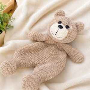 Könnte beinhalten: Ein hellbrauner Häkel-Teddybär als Schmusetuch mit weicher, strukturierter Garnoberfläche. Das Plüschtier hat einen runden Kopf mit weißer Schnauze, schwarzen Kunststoffaugen, einer schwarzen Nase und einem gestickten Mund an einem flachen Körper.