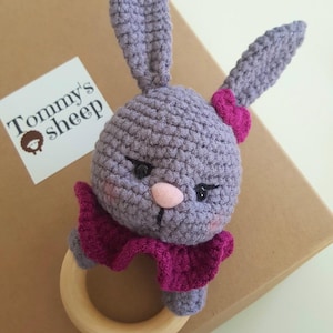 May include: A purple crocheted bunny rattle with a pink flower on its ear and a wooden ring. The bunny has black eyes and a pink nose.