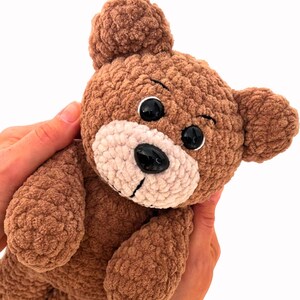 Puede incluir: Un oso de peluche de crochet marr&oacute;n con ojos negros y una nariz rosa. El oso est&aacute; siendo sostenido por las manos de una persona.