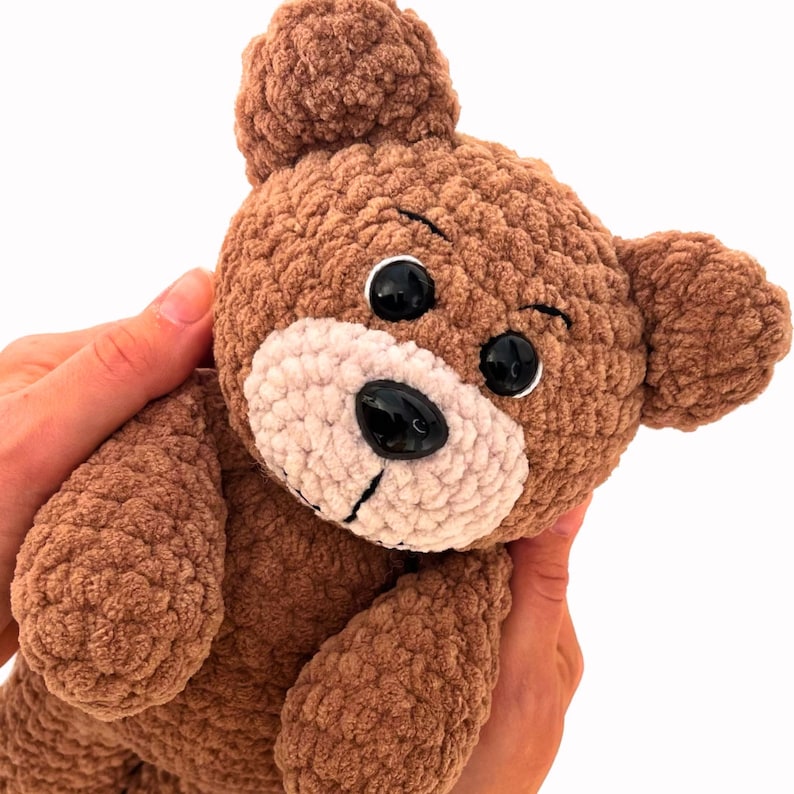 Puede incluir: Un oso de peluche de crochet marr&oacute;n con ojos negros y una nariz rosa. El oso est&aacute; siendo sostenido por las manos de una persona.