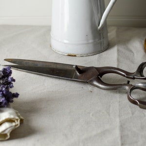 May include: A pair of antique shears with a long, curved blade and a wooden handle. The shears are lying on a white linen tablecloth.